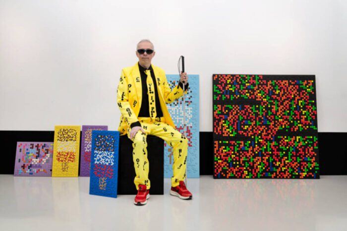 Artist, Clarke Reynolds, sitting on a box next to his braille tactile artwork. Clarke is wearing a yellow suit covered in black braille dots.