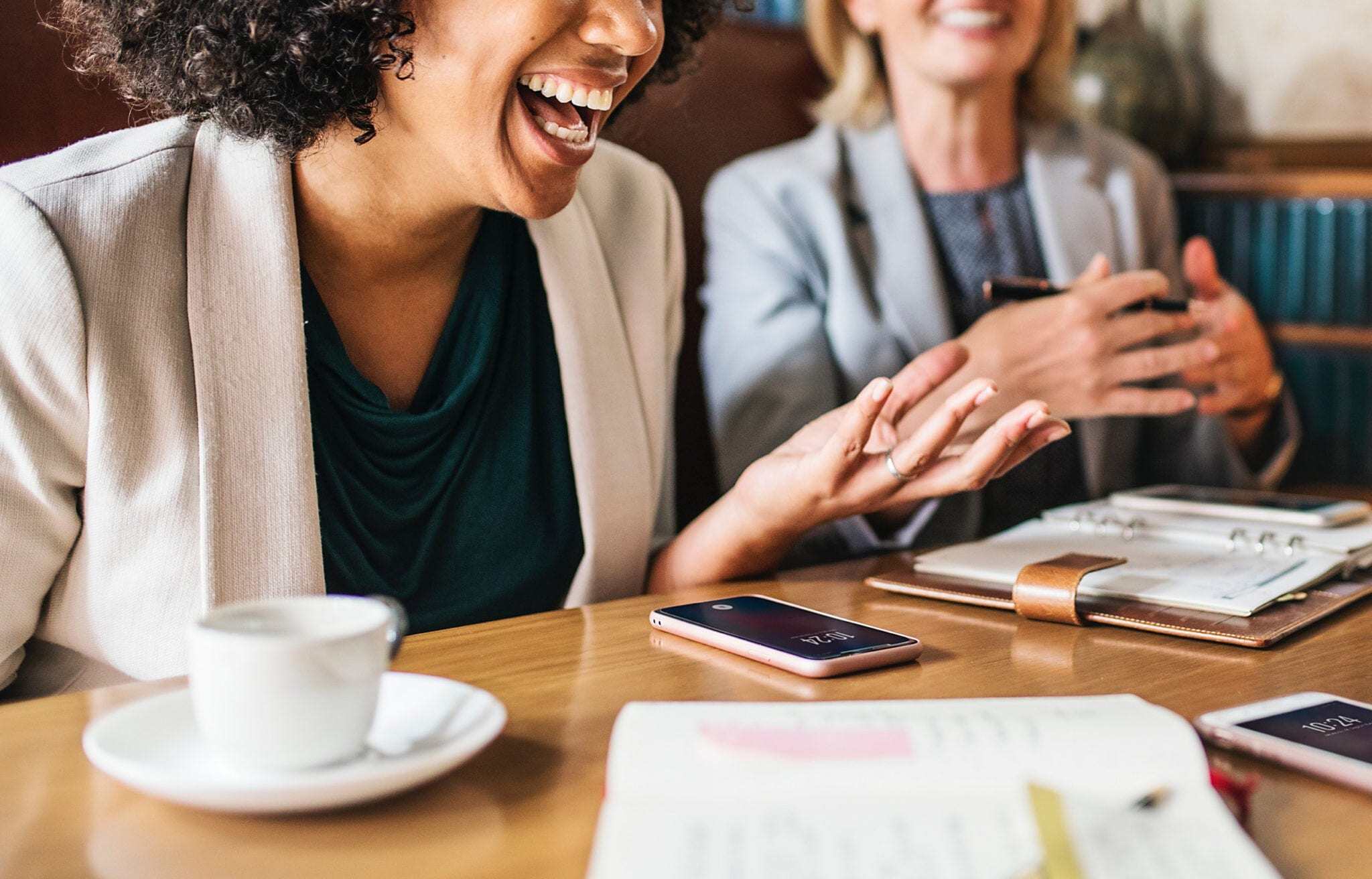 Women laughing while sitting at a table with a cup of coffee, phone and notebooks