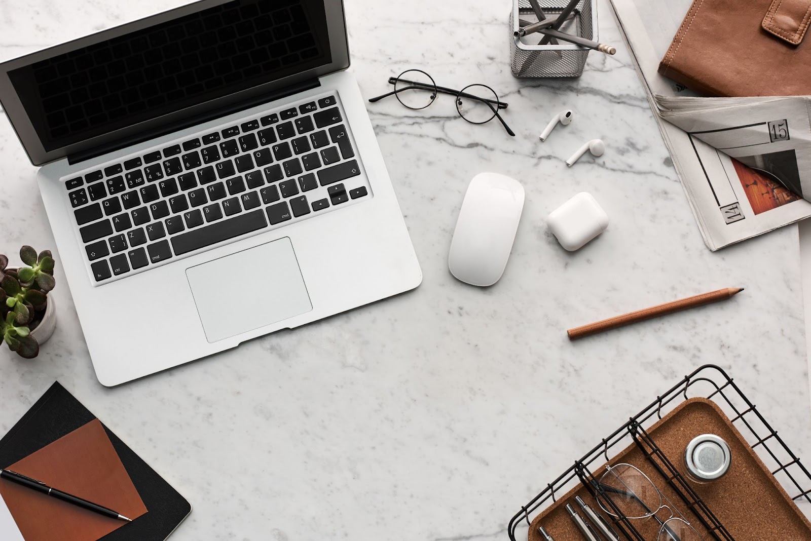 A laptop, computer mouse, a pair of air pods, a pair of reading glasses, and a pencil all sprawled out across a marble desk. 