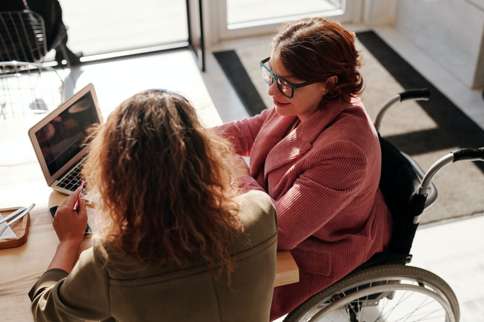 A women in a wheelchair looking at a computer screen with another women sitting next to her.